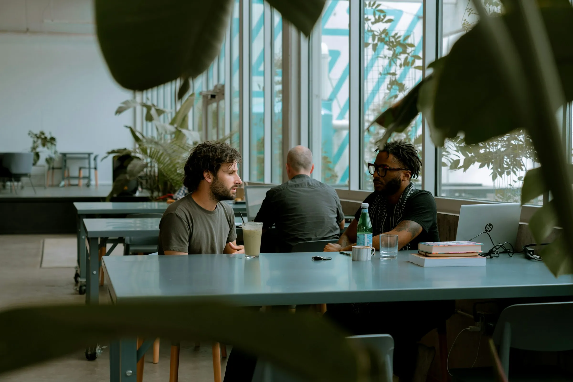 Three colleagues talk at a long table in a bright office with tall windows and plants
